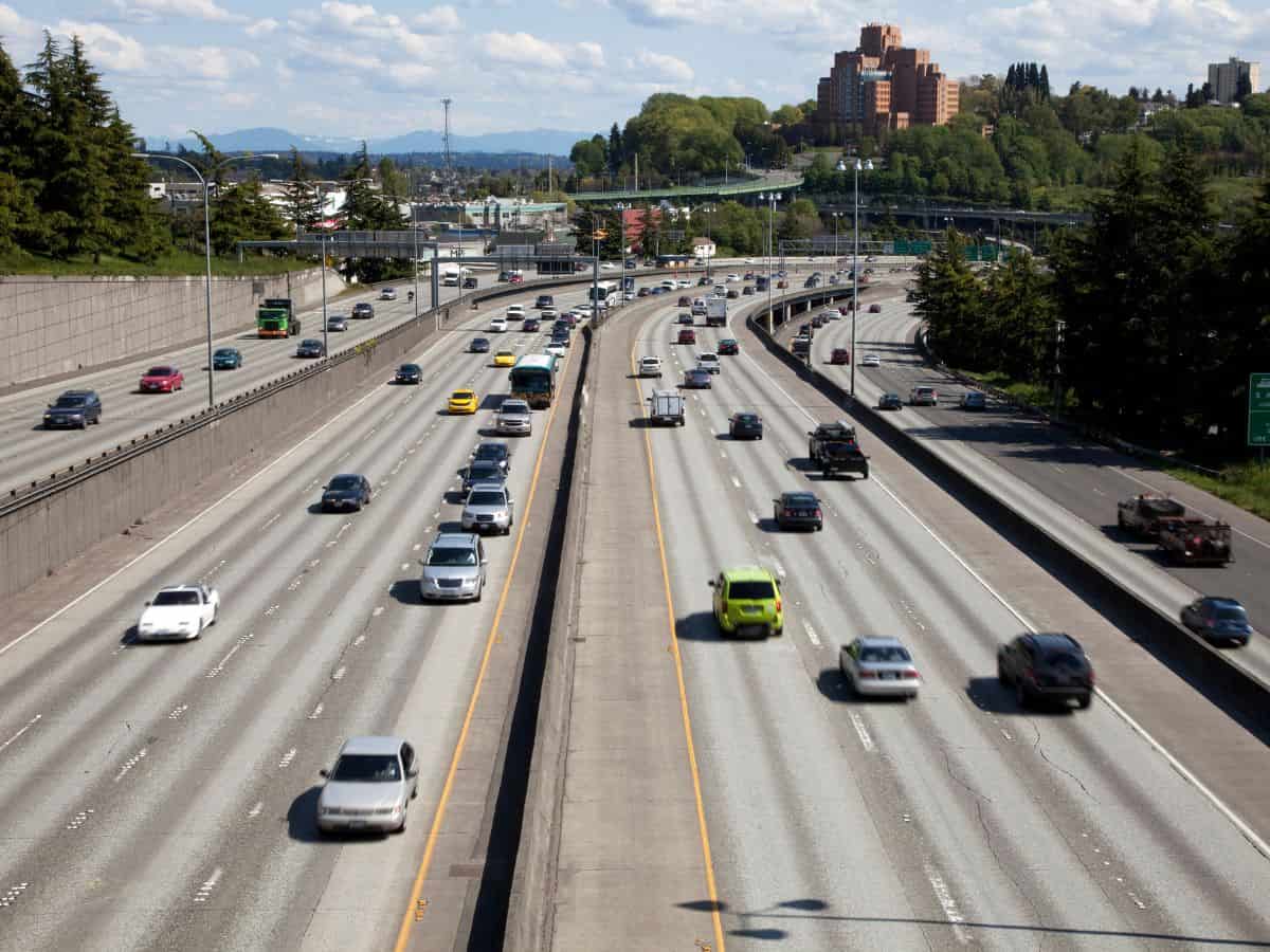 A multi-lane freeway in Seattle, showing several cars driving in both directions, with green trees and buildings in the background under a blue sky. This image illustrates the traffic conditions in Seattle vs. San Francisco, focusing on urban commuting and roadway infrastructure.