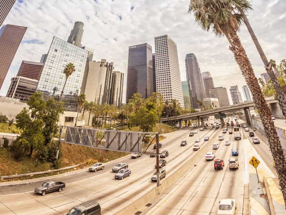 A busy Los Angeles freeway cutting through the downtown area, with tall palm trees and modern skyscrapers on both sides. Cars stream through the highway, showcasing Los Angeles' notorious traffic, making for an interesting comparison with New York City's subway-centric public transportation system.