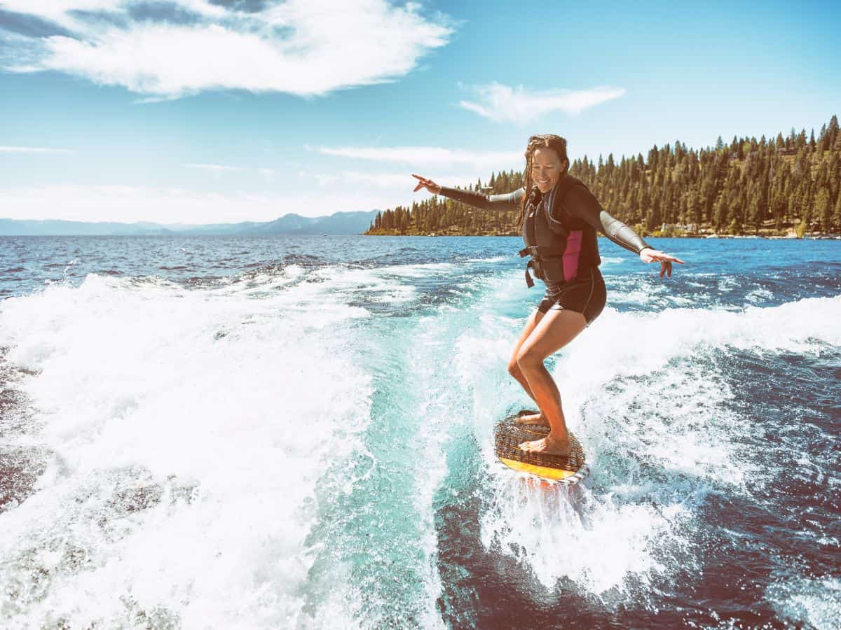 A woman enjoying a surfing experience on a lake, surrounded by a stunning natural backdrop of mountains and forests under a clear blue sky. This vacation scene captures the adventurous lifestyle one might find around both Seattle vs San Francisco, highlighting outdoor activities like water sports.
