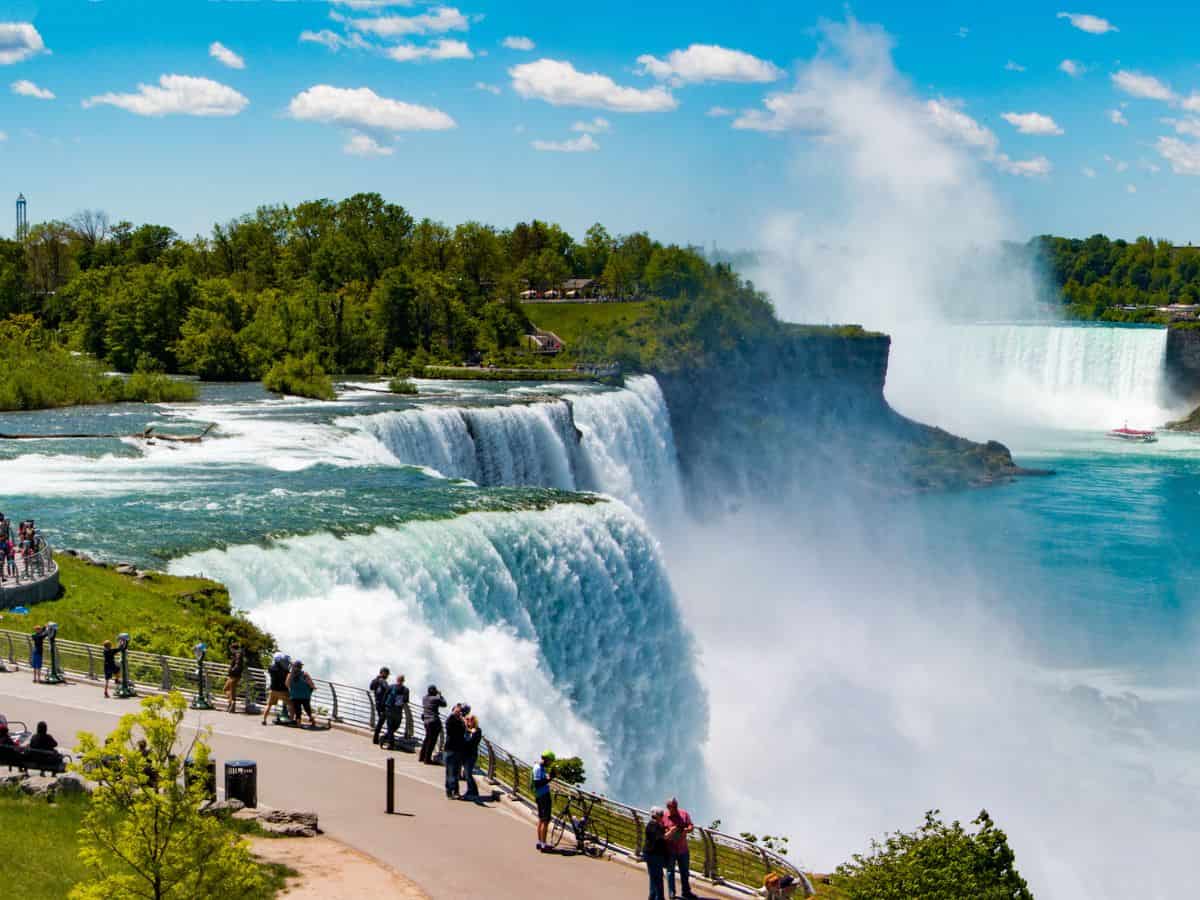 A picturesque view of Niagara Falls, with visitors admiring the natural wonder from an observation deck. The bright blue sky and vibrant greenery highlight the beauty of outdoor escapes available when comparing vacation spots linked to both coasts in Los Angeles vs New York
