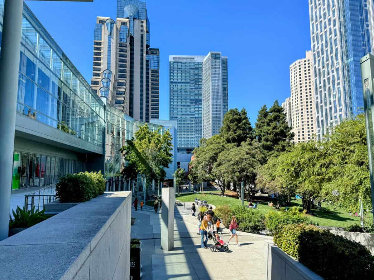 A modern urban plaza in San Francisco with glass buildings, lush greenery, and people enjoying the outdoor space on a sunny day. This image reflects the city's balance of nature and architecture, contributing to the discussion of weather and outdoor living in the "San Francisco vs Los Angeles" comparison.