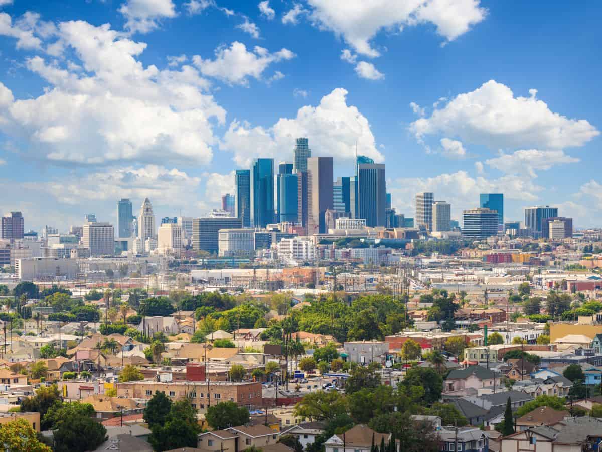 A panoramic view of a sprawling urban area with a densely packed skyline under a bright blue sky filled with fluffy clouds. The scene captures the contrast between residential areas and high-rise buildings, representing weather conditions and cityscapes in the Seattle vs. San Francisco comparison. The clear, sunny weather indicates the diversity in climate between these cities.
