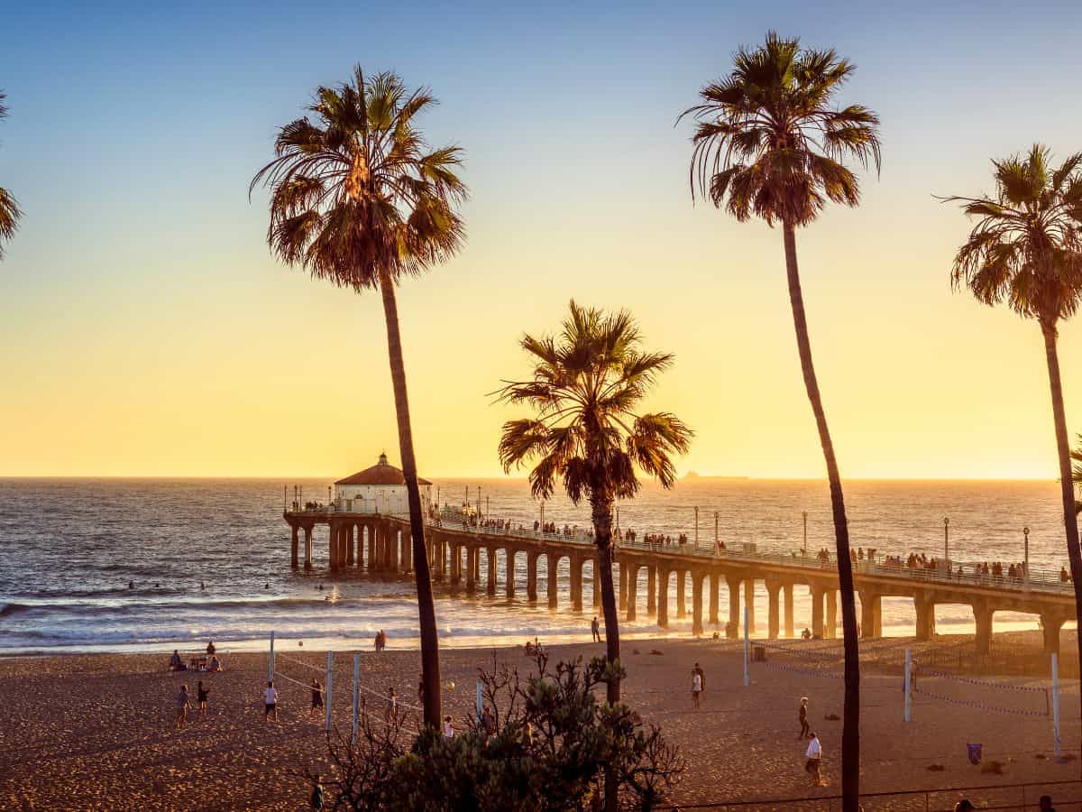 A stunning beach scene at sunset in Los Angeles, with the iconic Manhattan Beach pier stretching into the Pacific Ocean. Towering palm trees frame the beach, where people are enjoying a game of volleyball. The warm golden glow of the setting sun contrasts the weather in Los Angeles vs New York City, highlighting LA's sunny and laid-back vibes.