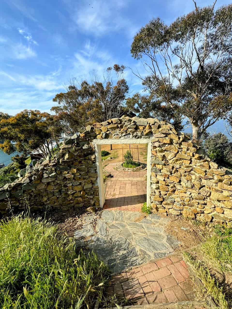 A picturesque doorway made of stone with a path leading to a garden, overlooking a hilly and vibrant green landscape under a bright blue sky.