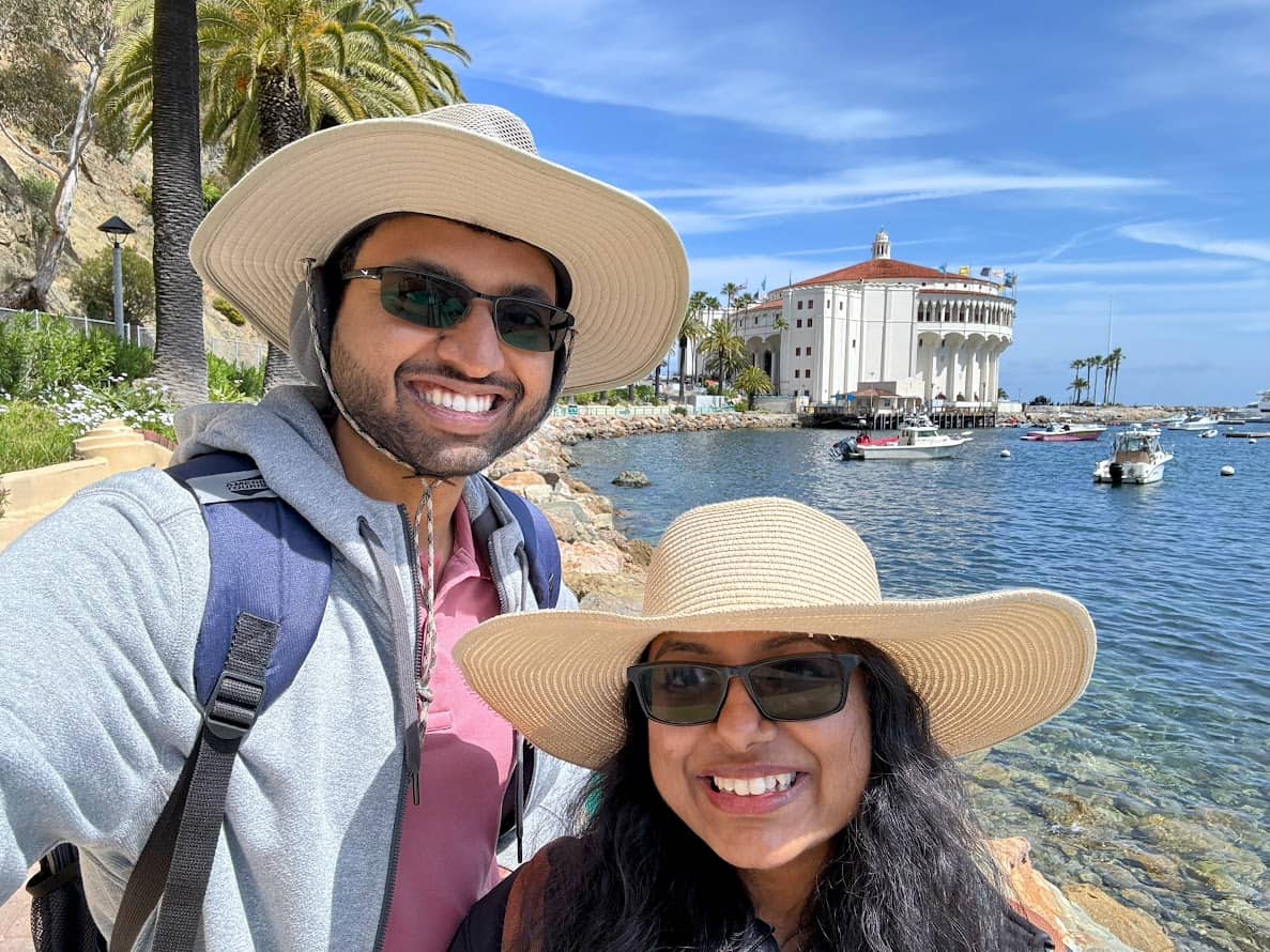 Kiran and Shreeyeh wearing wide-brimmed hats smiling in front of the Avalon harbor, with the iconic Casino building and palm trees visible in the background.