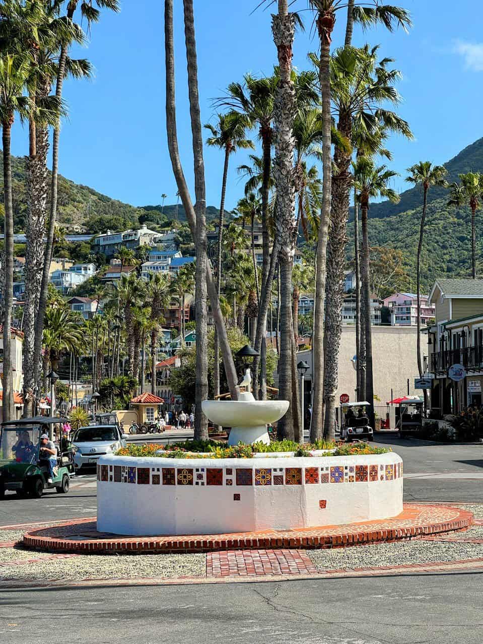 A white tiled fountain decorated with colorful mosaic tiles, surrounded by tall palm trees and small buildings in the heart of Avalon.