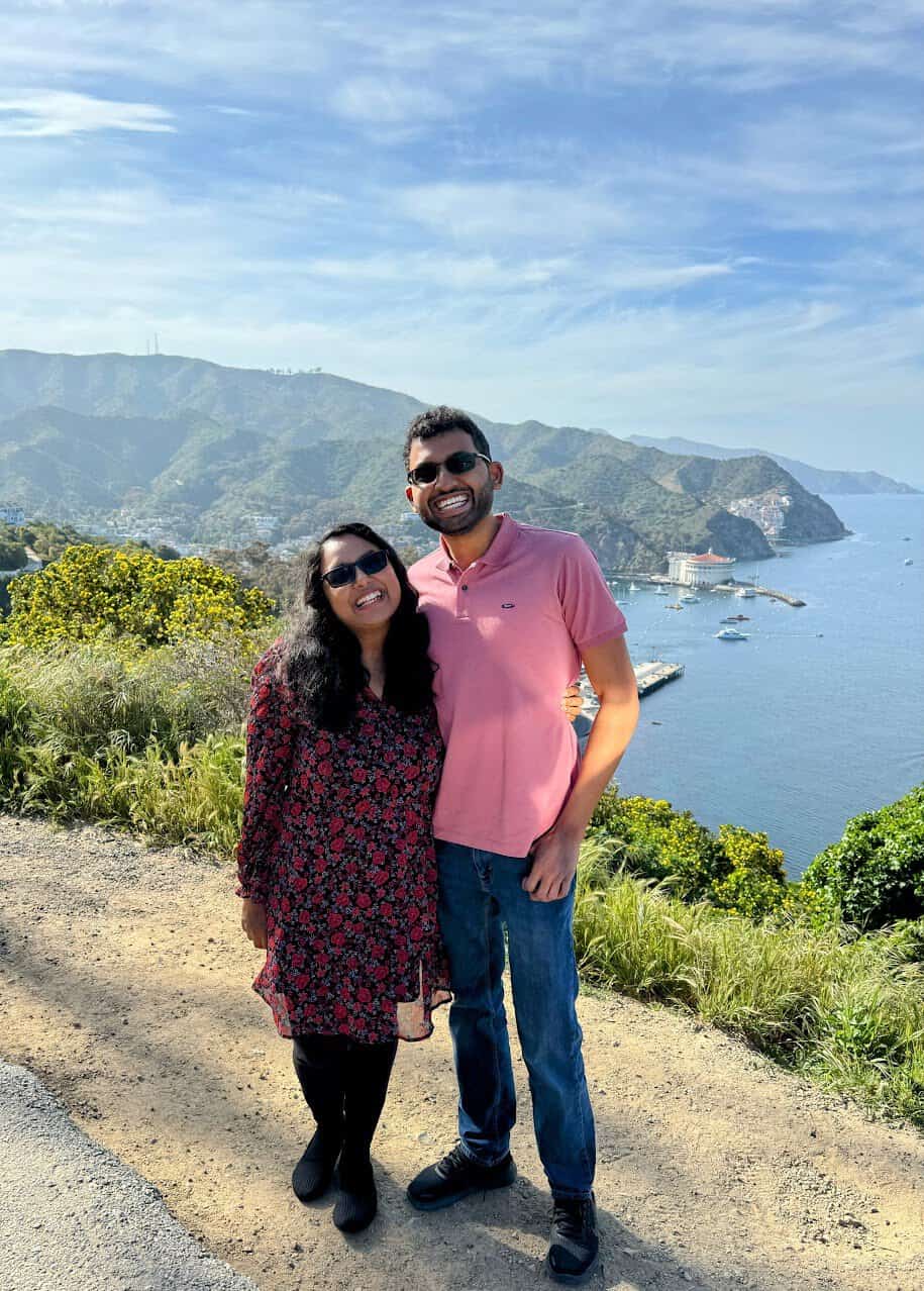 Shreeyeh and Kiran standing on a hill with the scenic Avalon harbor and hilly terrain in the background under a sunny blue sky.