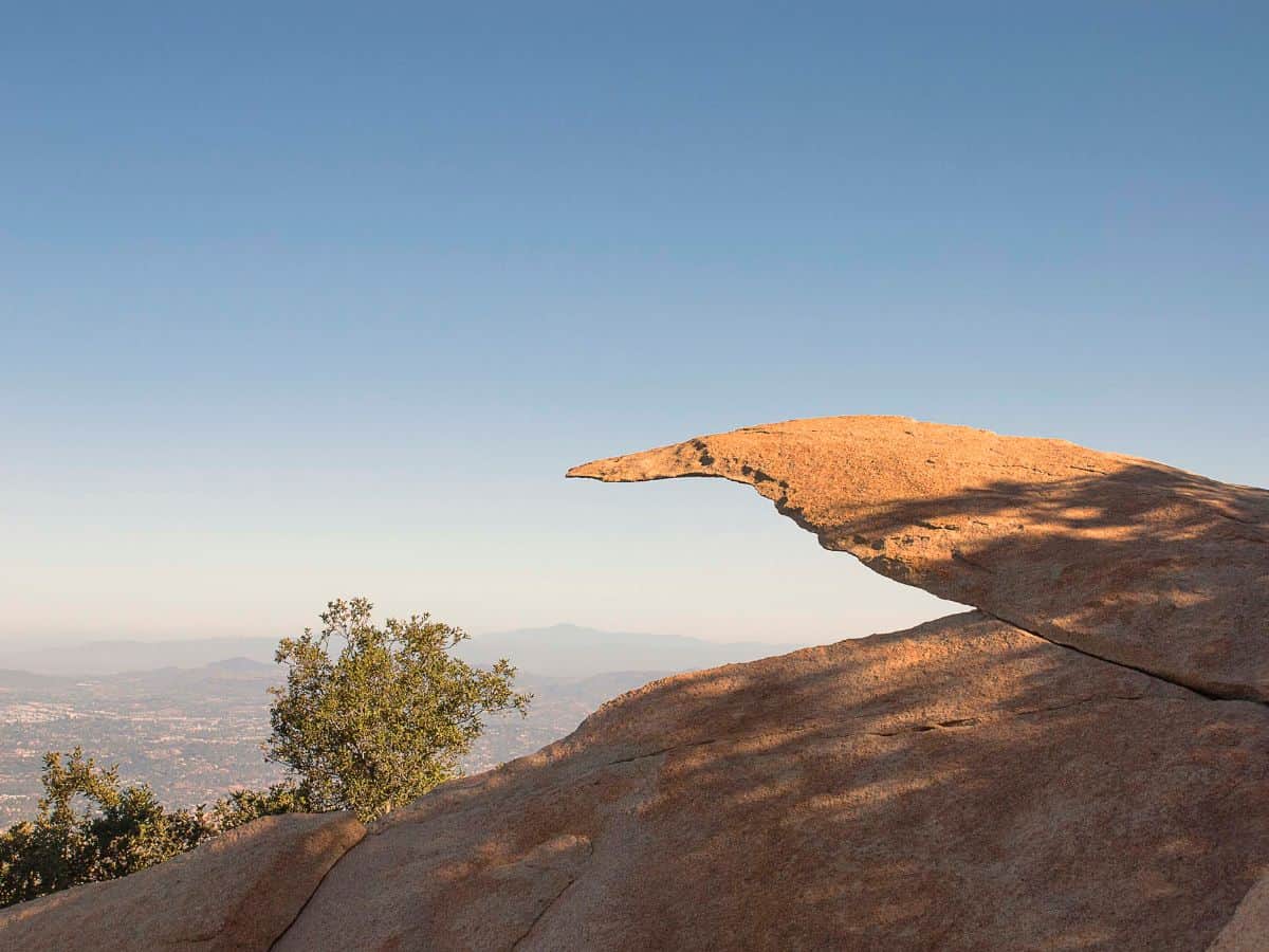 A close-up of the famous Potato Chip Rock formation on Mount Woodson Trail, perched against a backdrop of clear skies and distant mountains.