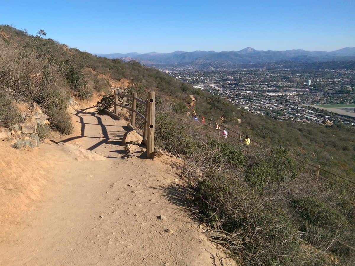 A scenic trail at Cowles Mountain with a dirt path leading uphill, bordered by shrubs and a wooden fence, offering panoramic views of San Diego in the distance, ideal for dog-friendly hikes in San Diego.