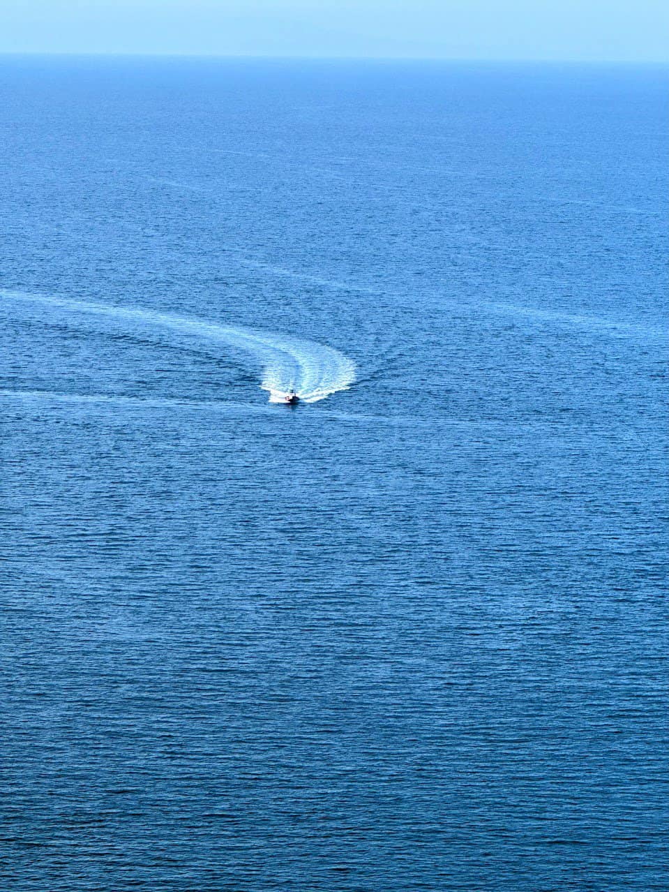 A boat creating a gentle wake on the deep blue ocean, heading towards the horizon under a bright and clear sky.