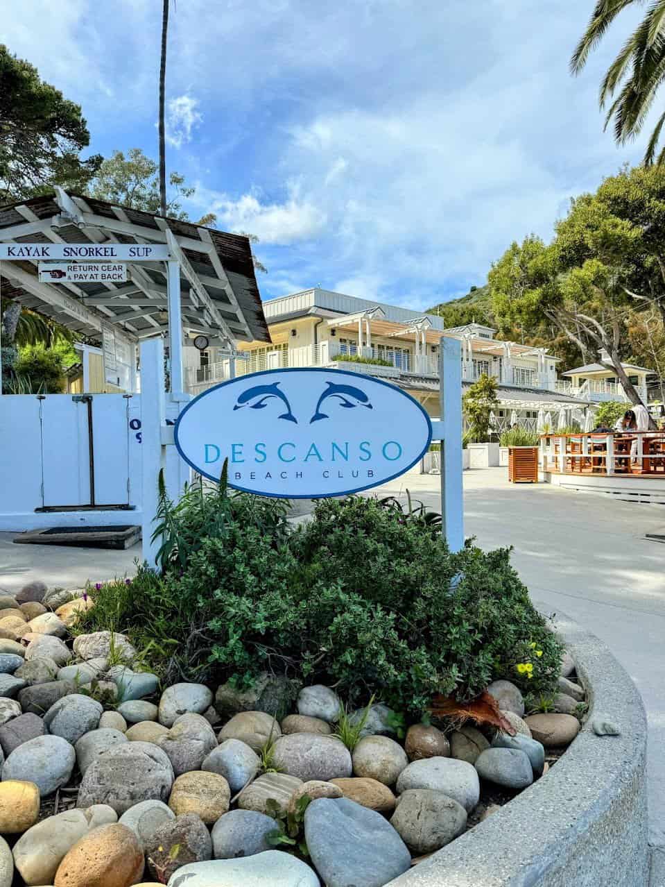A welcoming sign for Descanso Beach Club surrounded by neatly arranged rocks and vibrant greenery under a clear sky, with the club's white buildings visible in the background.