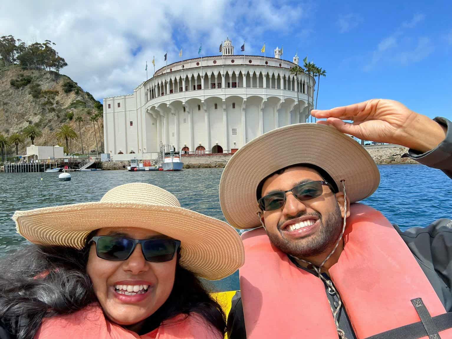 Shreeyeh and Kiran in sun hats and life vests smiling on a kayak, with the iconic white Avalon Casino building and palm trees in the background, framed by blue skies and the sparkling ocean.
