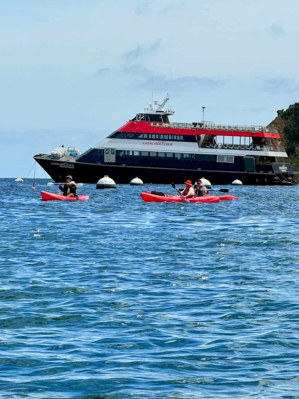 A group of kayakers paddling in the calm blue waters near Catalina Island, with the large red and black Catalina Flyer ferry anchored nearby.