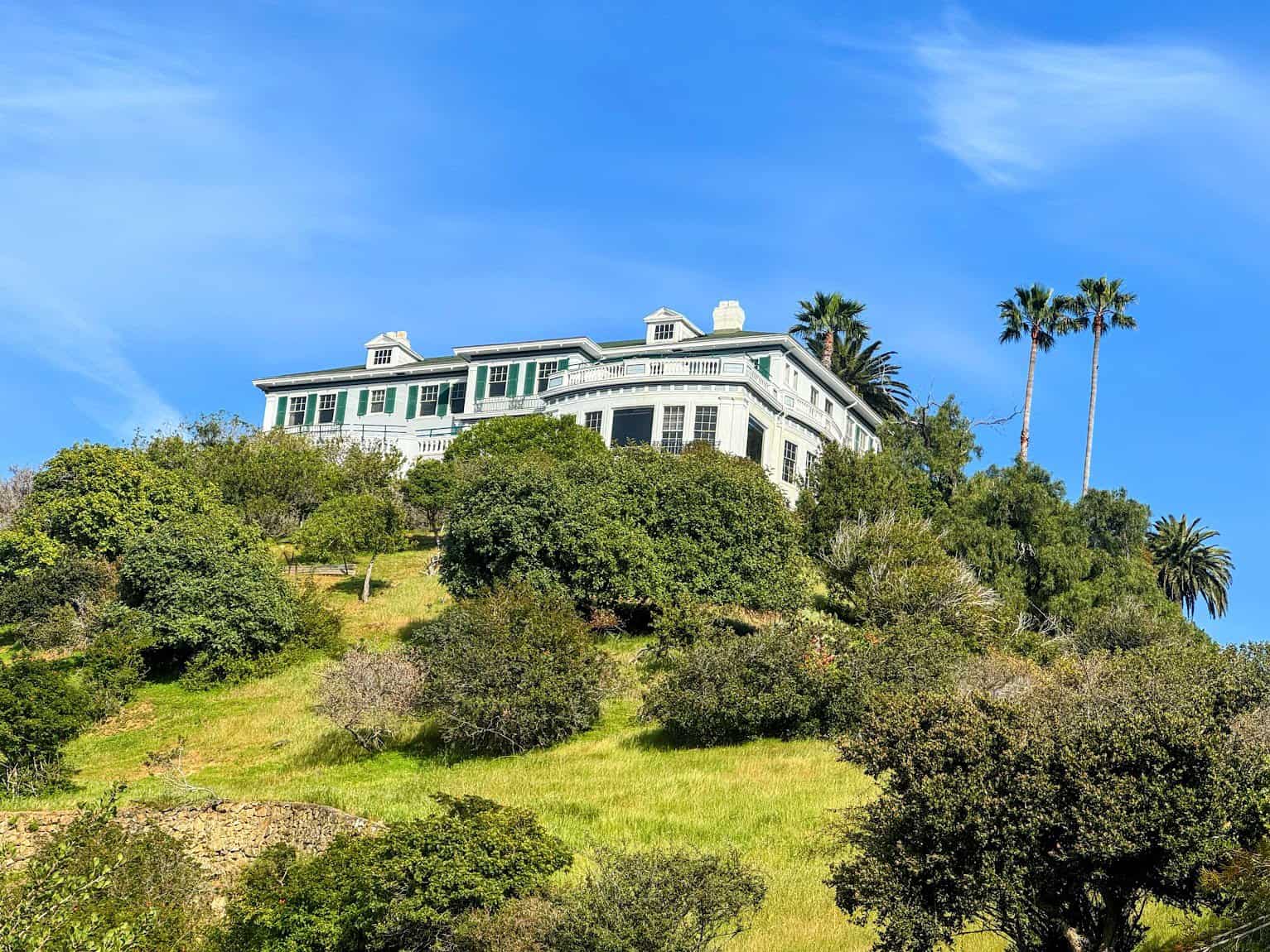 A grand white colonial-style building known as Mt. Ada, perched on a green hillside surrounded by lush trees and tall palm trees, set against a bright blue sky.
