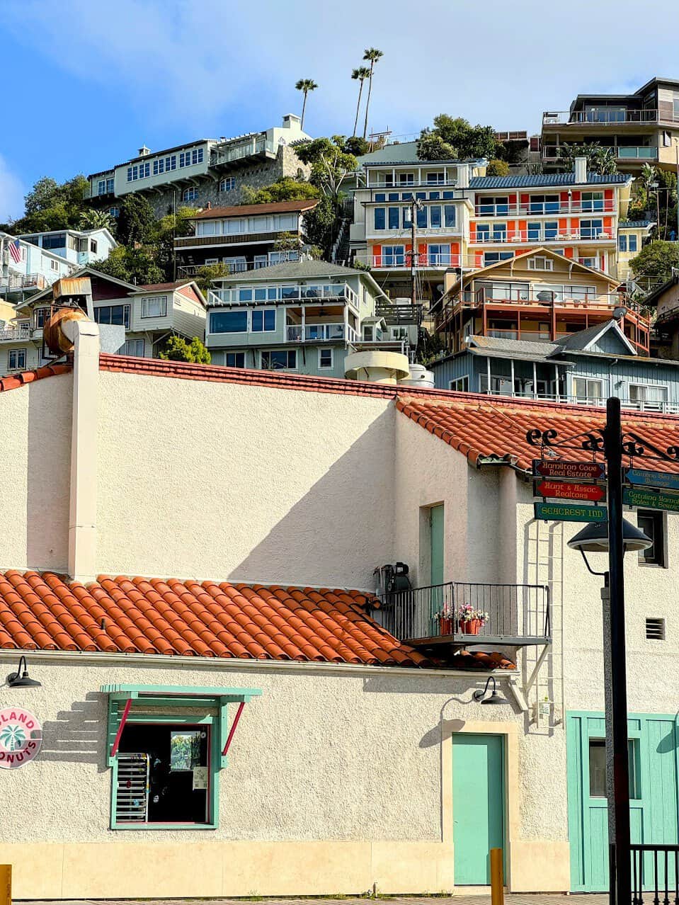 A front-facing view of a small, white building with a tiled red roof, situated at the base of a hill filled with multicolored homes stacked vertically along the slope. A donut shop window adds a quaint charm.