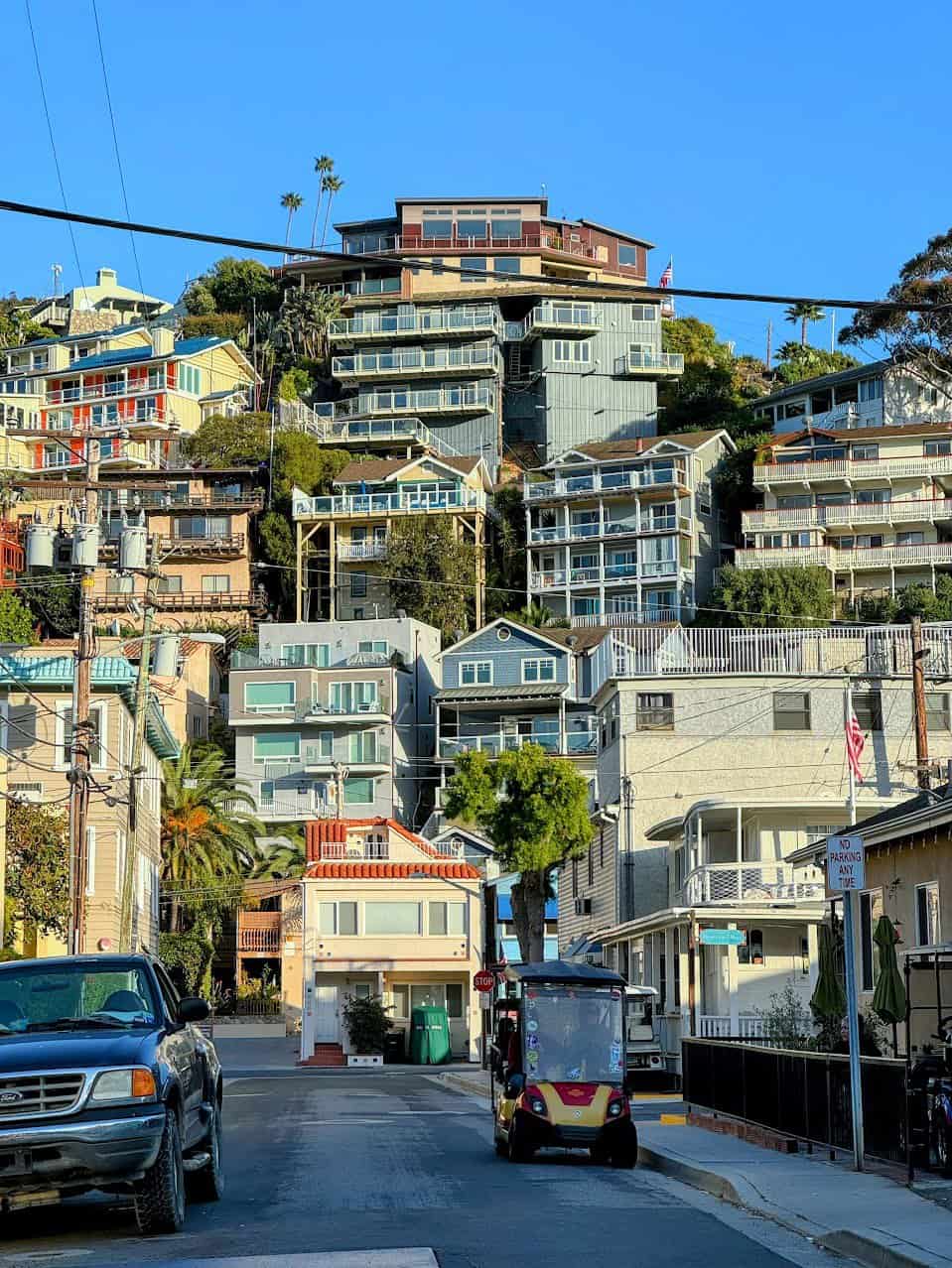 A lively street view of Catalina Island showcasing stacked hillside houses in various colors, with a small golf cart parked in the foreground under the clear blue sky.