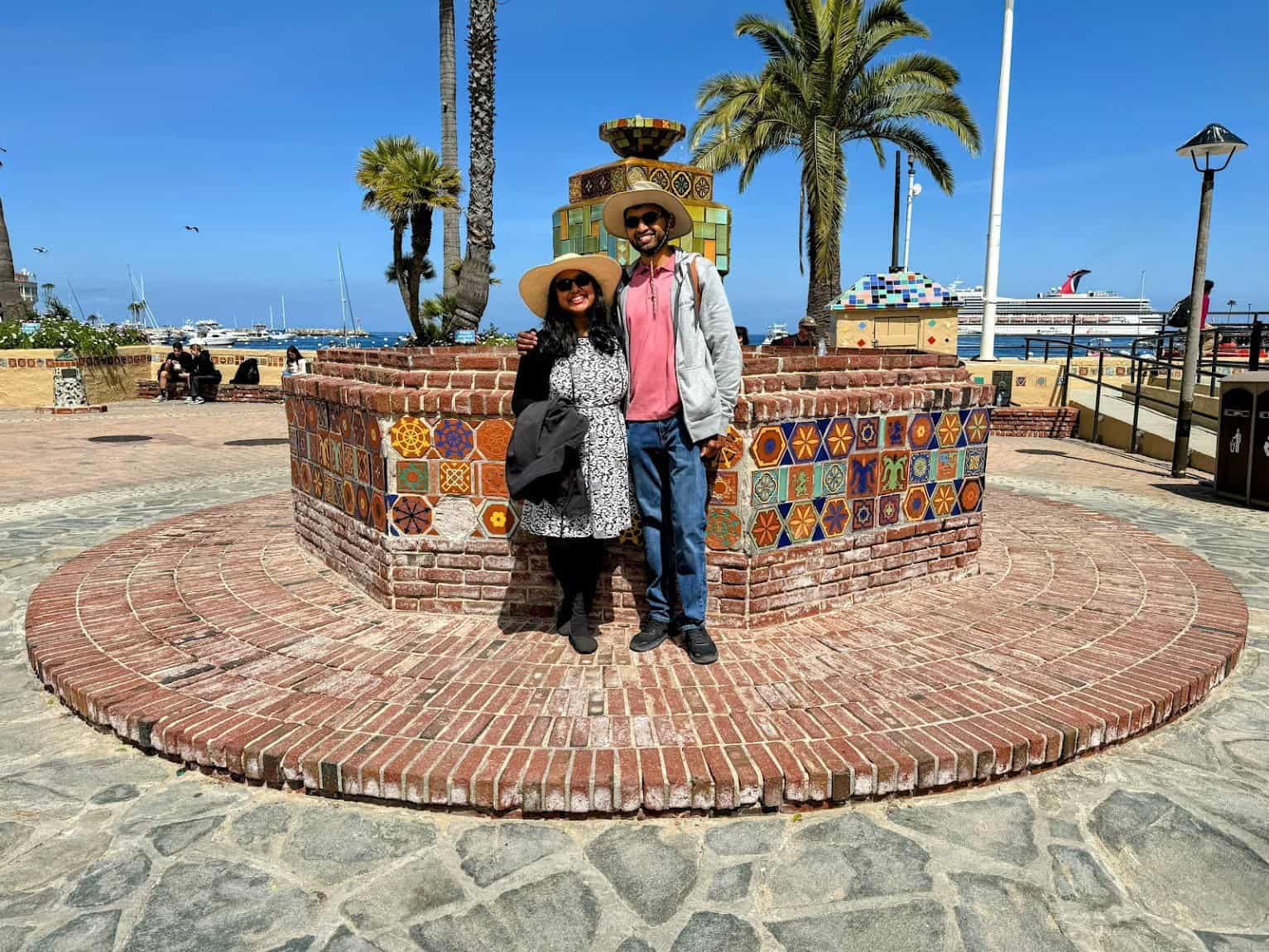 Shreeyeh and Kiran posing in front of the Wrigley Fountain on Catalina Island, a colorful tiled circular fountain surrounded by palm trees. The backdrop includes a clear blue sky, the harbor with yachts, and a large cruise ship docked in the distance.