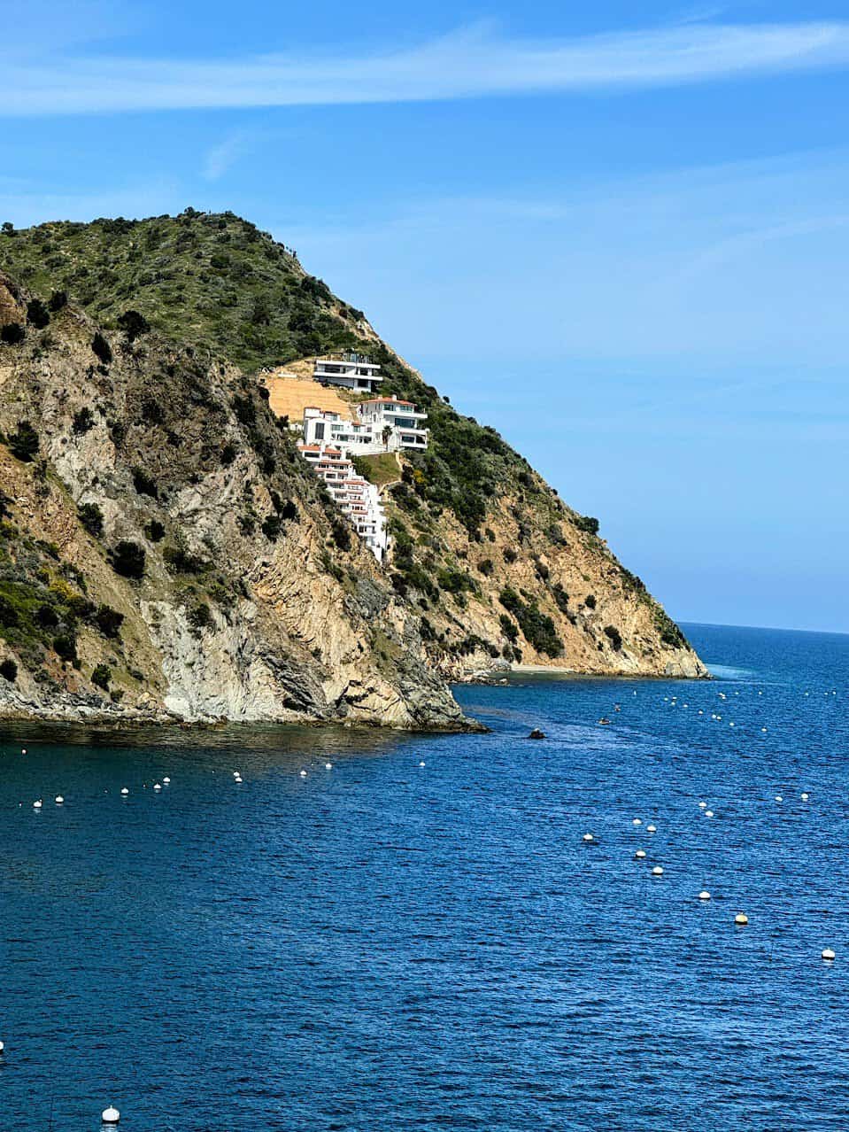  A striking modern home built into the side of a steep rocky cliff on Catalina Island, overlooking a pristine blue ocean dotted with moored buoys.