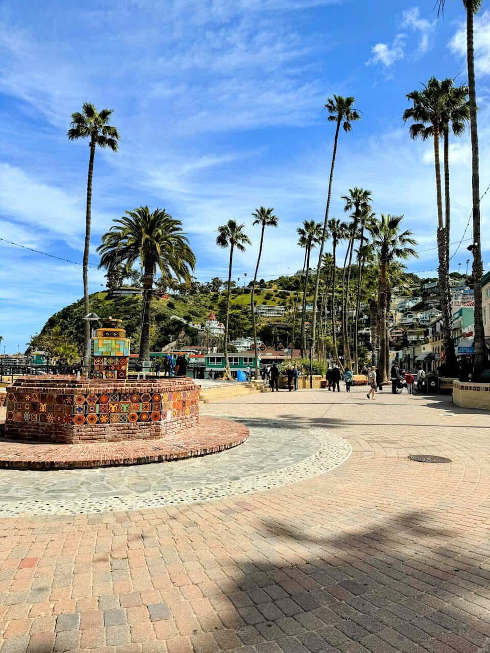 A vibrant, tiled fountain surrounded by palm trees under a bright blue sky, located in Avalon on Catalina Island. The fountain serves as a focal point in the charming plaza, with green hills and quaint buildings in the background.