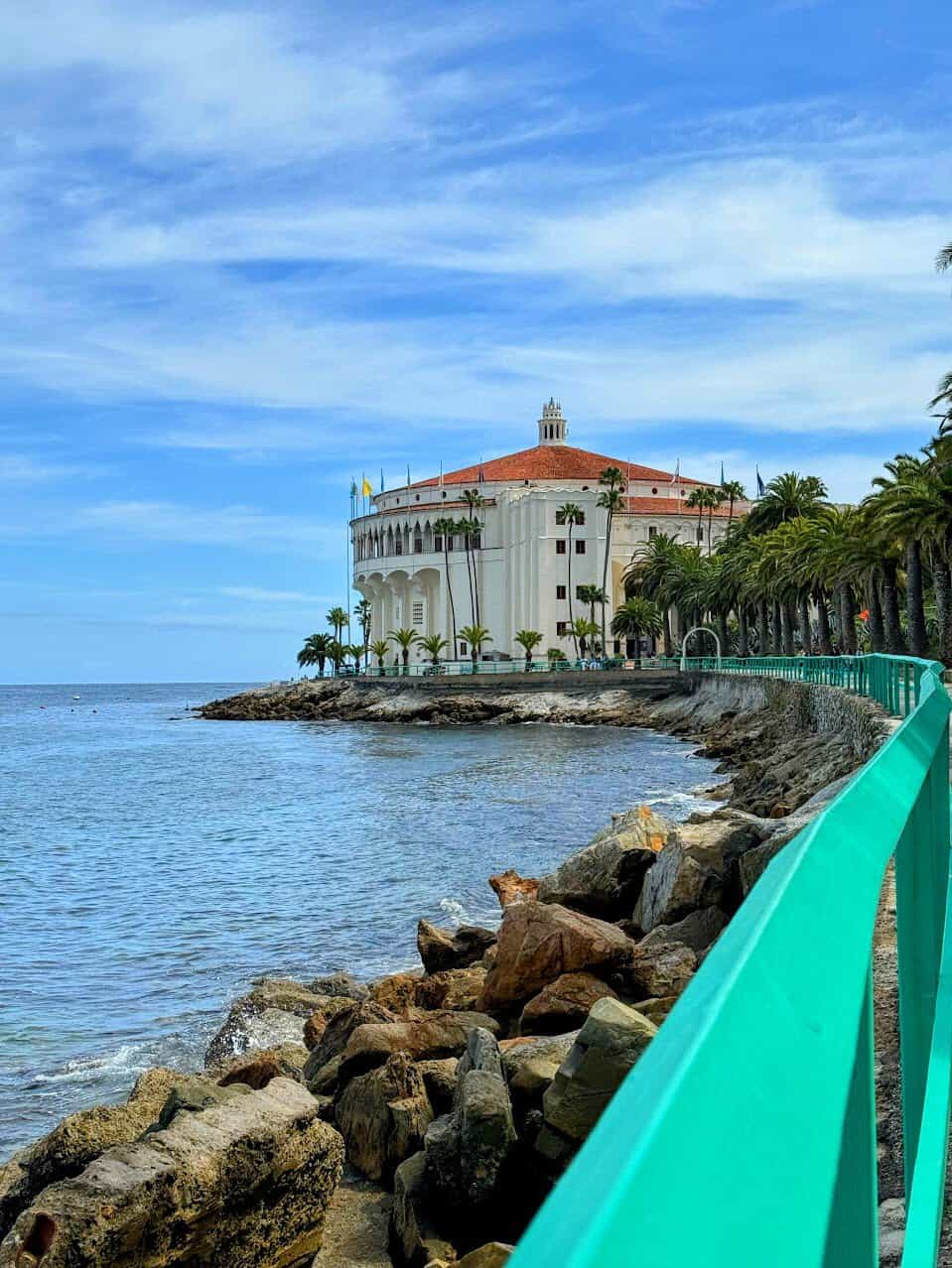 A view of the iconic Catalina Casino, a white building with a red roof and classic architecture, perched along the rocky shoreline of Catalina Island. A turquoise railing lines the foreground, with calm ocean waters and palm trees adding to the scenic beauty.