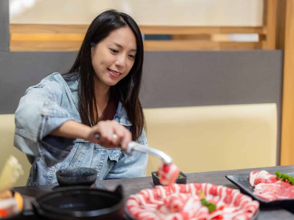 A woman wearing a denim jacket sitting at a table, enjoying a hot pot meal. She is using a tong to dip meat slices into a simmering pot. The atmosphere is cozy, evoking the authentic hot pot San Francisco dining experience.