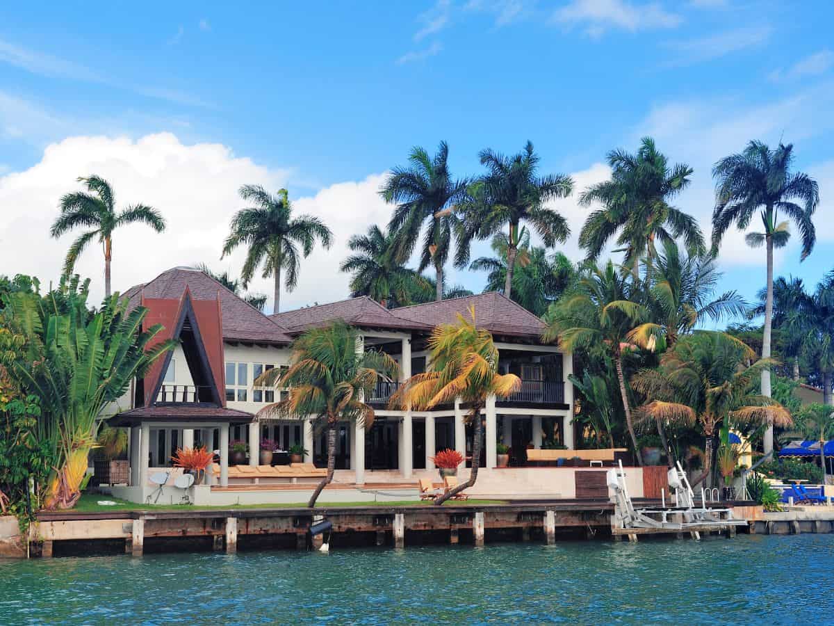  A luxurious beachfront house in Miami, surrounded by palm trees, facing the water. The house features modern architecture with large windows and a spacious patio. This image represents the style of high-end lodging commonly found in Miami vs Los Angeles.