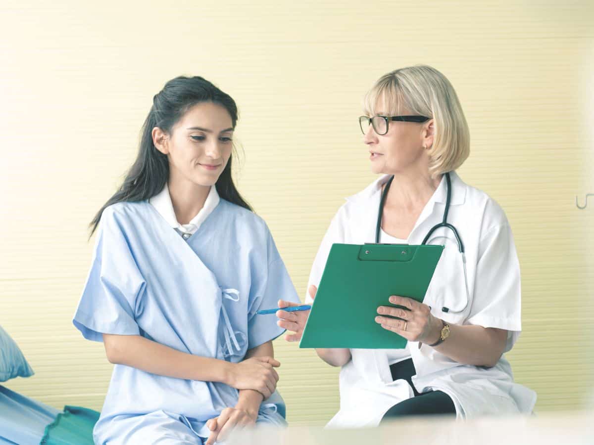 A doctor discussing a medical report with a female patient in a clinical setting. The patient is sitting in a hospital gown, indicating healthcare services available in Los Angeles, which can be compared with those in Miami.