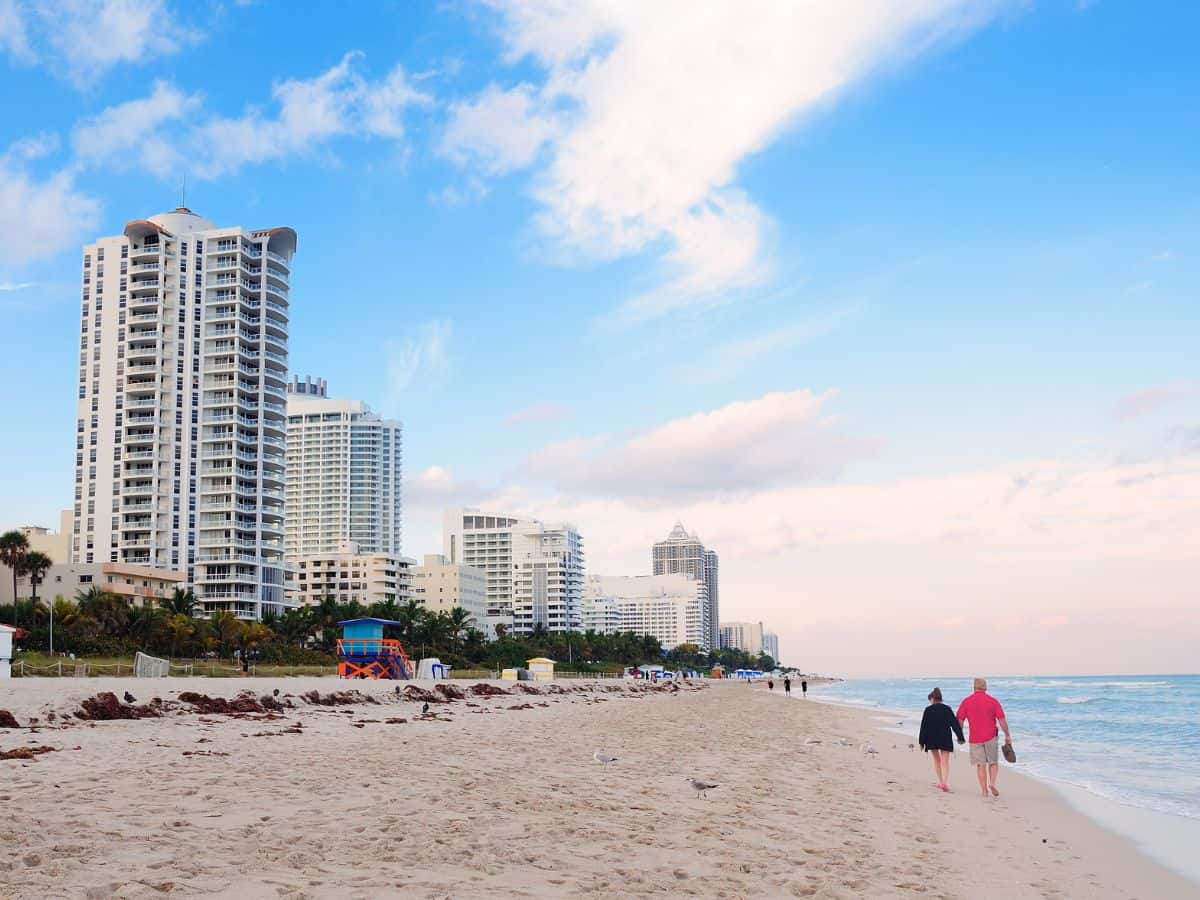  A sandy beach in Miami with high-rise buildings lining the shore. A couple is seen walking along the beach, highlighting the luxurious yet potentially expensive lifestyle, often associated with Miami vs Los Angeles.