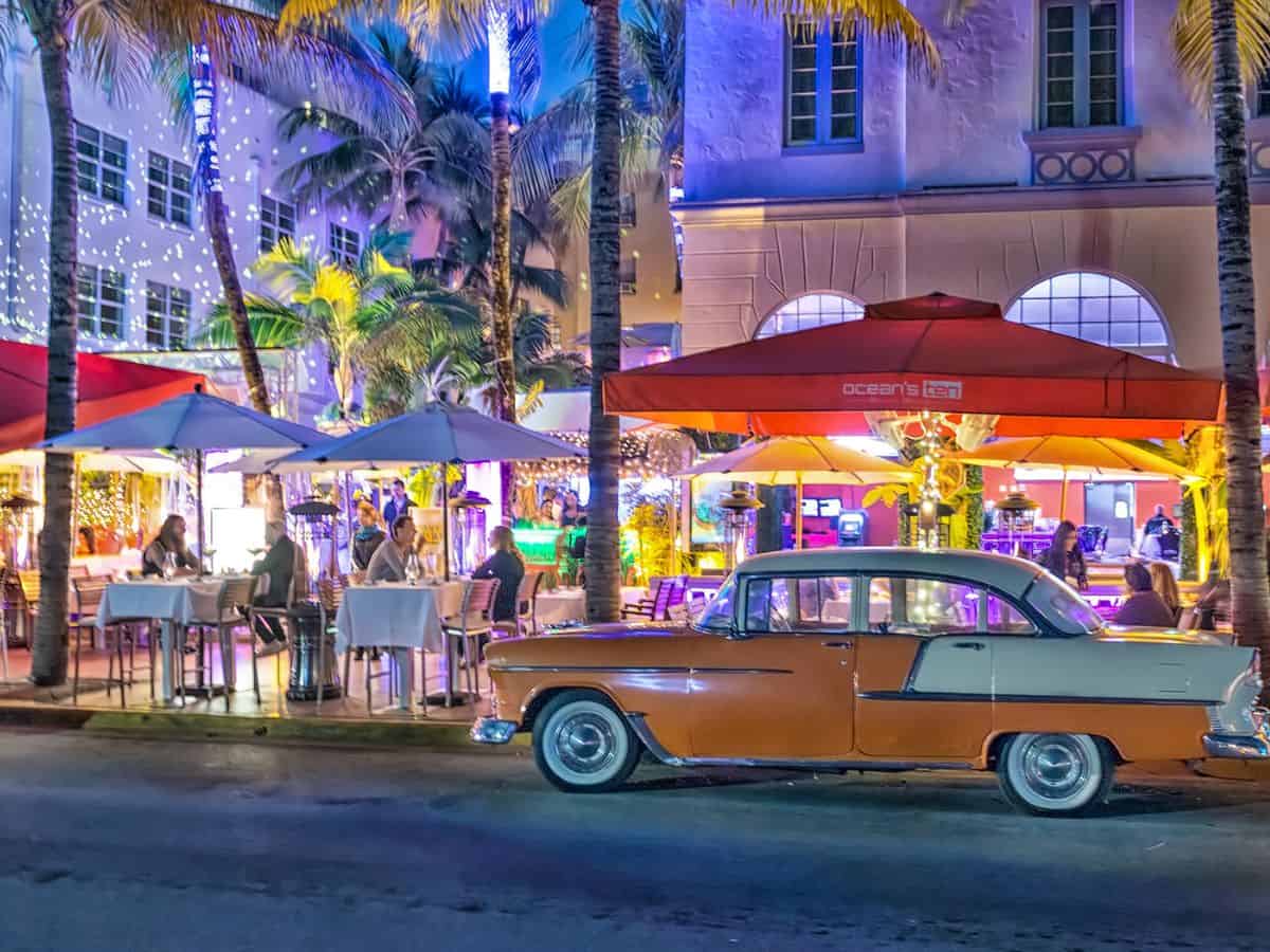 A vibrant nighttime scene on Ocean Drive in Miami Beach, featuring an outdoor dining area with colorful lights. A classic vintage car is parked in front, adding to the lively atmosphere that contrasts with the nightlife found in Los Angeles.