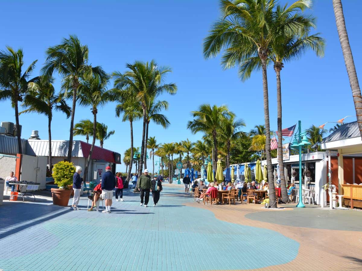 A bustling promenade in Miami, lined with palm trees and outdoor cafes. The scene shows people strolling and dining under a bright blue sky, representing Miami's lively population and outdoor lifestyle, a contrast to the urban density of Los Angeles in the Miami vs Los Angeles comparison.