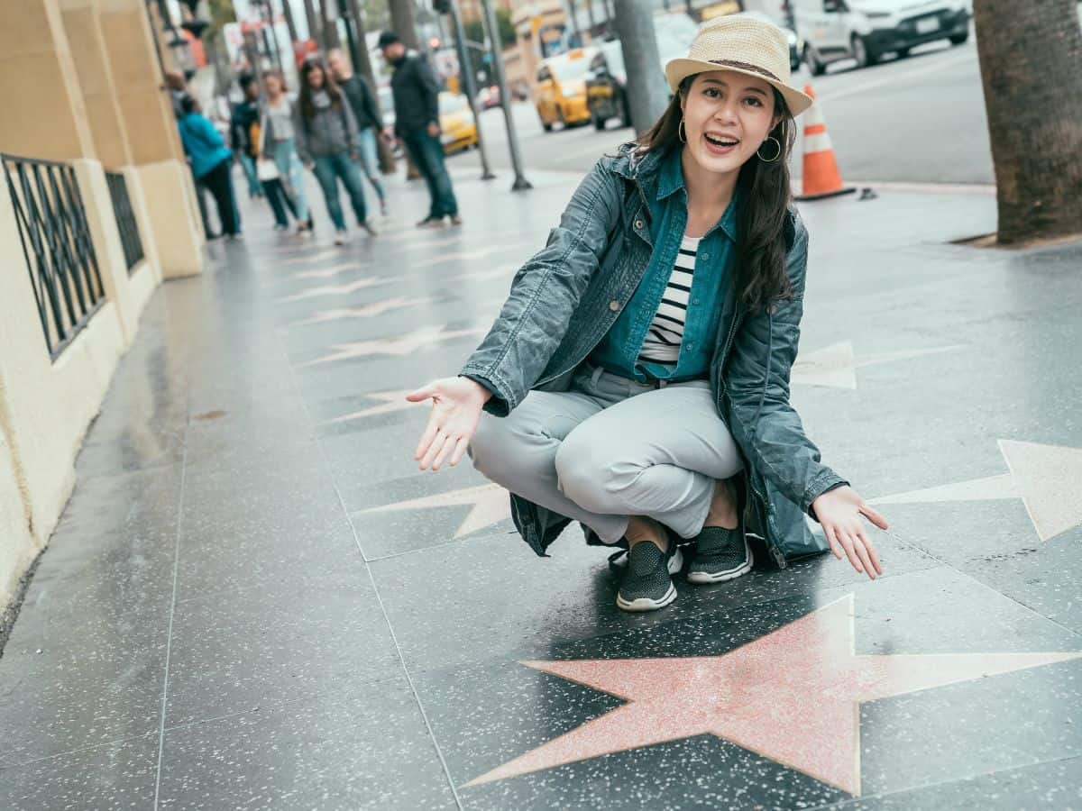 A woman excitedly pointing to a star on the Hollywood Walk of Fame in Los Angeles. The background shows other tourists and city life, representing aspects of the quality of life in Los Angeles vs Miami.
