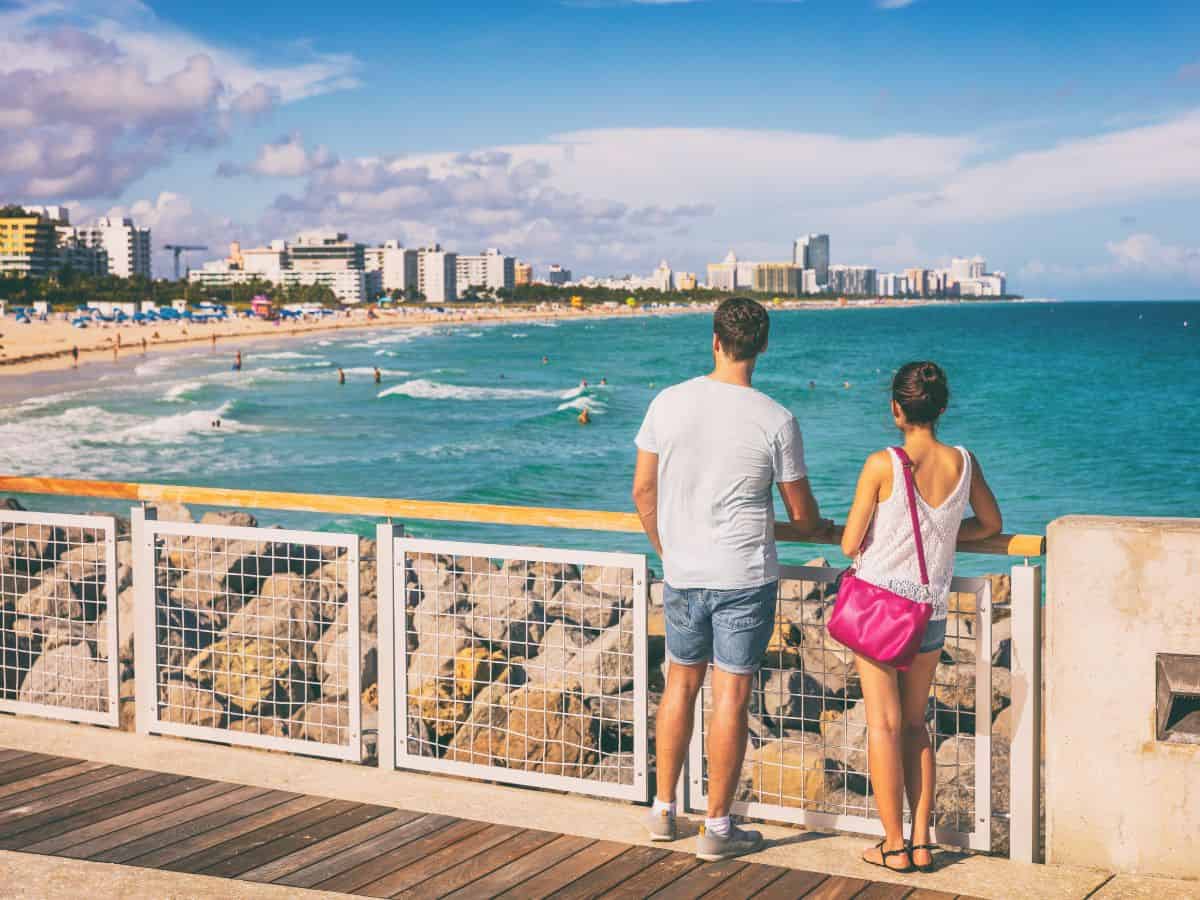 A couple standing on a boardwalk overlooking Miami Beach. The turquoise ocean and sandy shore stretch into the distance, with Miami's city skyline visible in the background. This scene captures the vacation vibe of Miami, in contrast to a similar experience in Los Angeles.