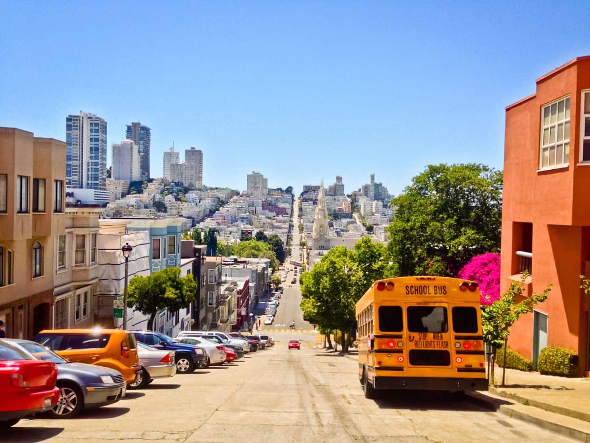 A view down a steep hill in San Francisco, with a school bus driving down and rows of parked cars. The background shows densely packed buildings leading toward the horizon, capturing the contrast in weather conditions between Denver vs San Francisco.
