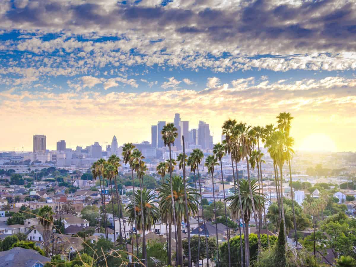  A panoramic view of downtown Los Angeles at sunrise, with palm trees in the foreground and a partly cloudy sky above. The image captures the warm, sunny climate characteristic of Los Angeles, providing a comparison point with Miami's tropical weather in the Miami vs Los Angeles debate.