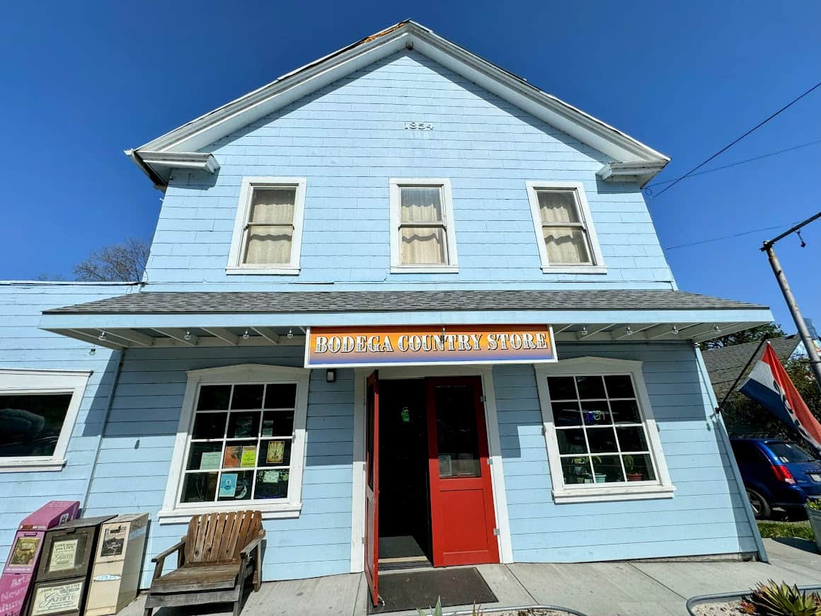 The charming blue exterior of Bodega Country Store with a red door, white trim, and wooden chairs on the front porch, under a clear blue sky with signs for local newspapers and vintage advertisements in the windows.
