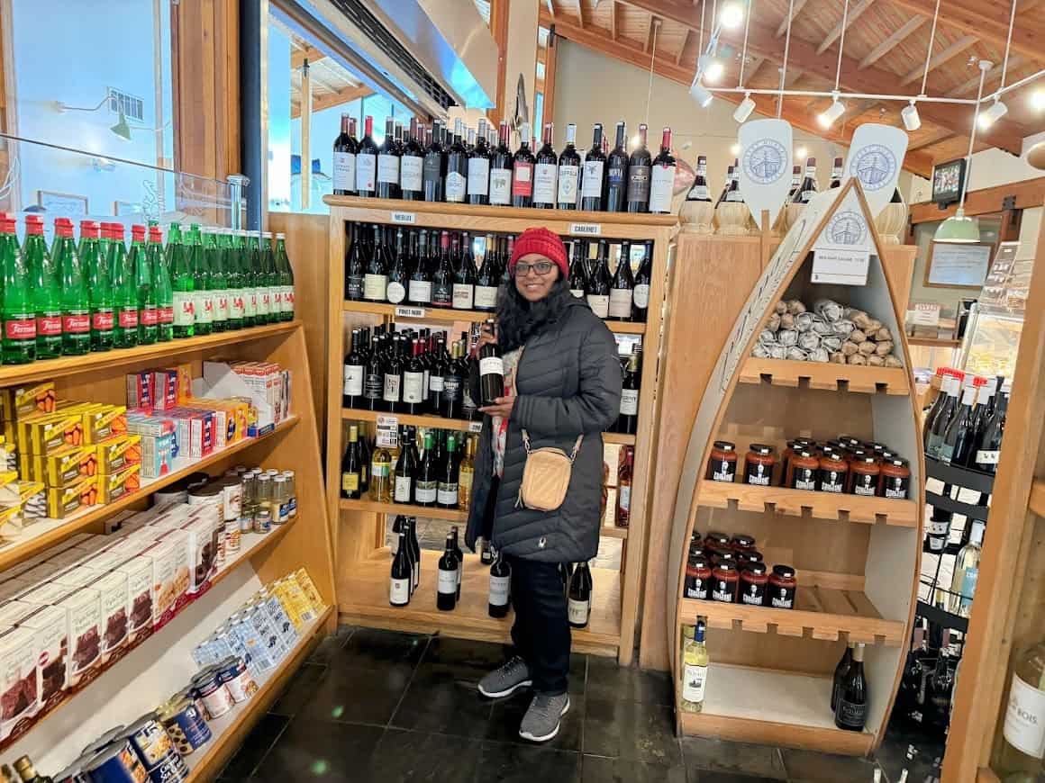 A warmly lit gourmet grocery store with shelves of wine, sparkling water, and artisanal goods, featuring a woman in a winter coat holding a wine bottle while browsing the wine selection.