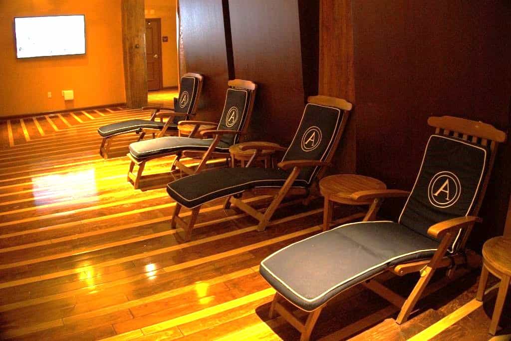 A row of dark wooden deck chairs with navy blue cushions, each bearing a circular 'A' emblem, arranged on a patterned wooden floor, facing a television mounted on the wall.