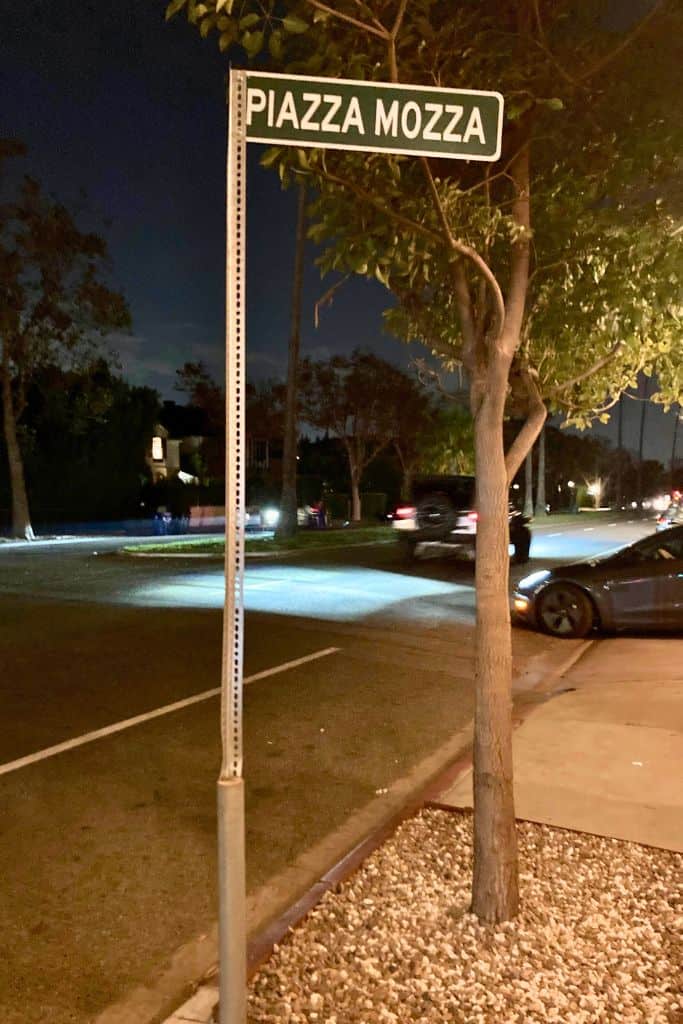 A street sign at night that reads 'PIAZZA MOZZA' with a tree next to it, and cars parked in the background under streetlights.