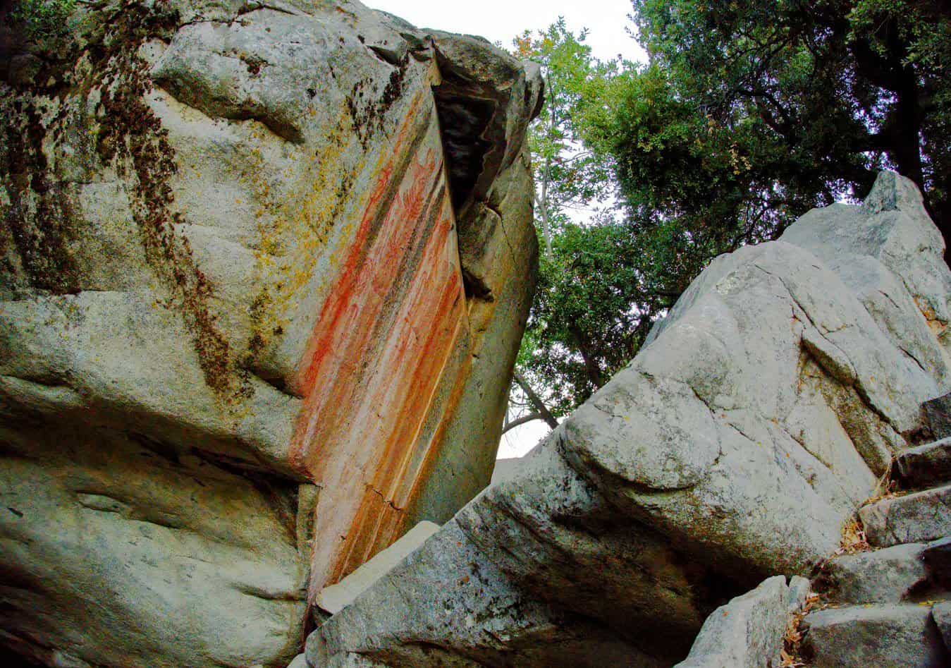 A large rock with a naturally eroded vertical face, displaying pictographs set against other massive boulders and foliage, indicative of a rugged terrain.