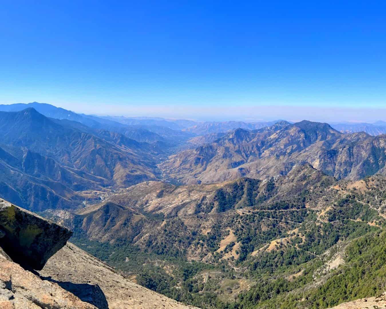 A breathtaking view from Moro Rock in Sequoia National Park, showcasing a vast expanse of rugged mountain ranges and valleys under a clear blue sky. The foreground features a rocky outcrop, while the landscape reveals a mix of green forested areas and dry terrain stretching into the distance.