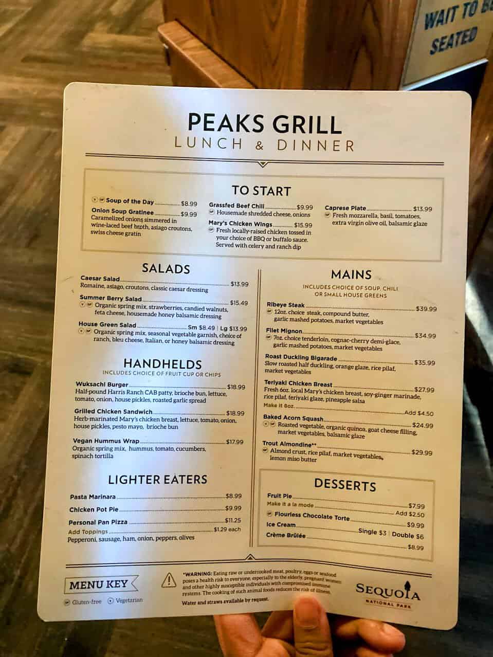 A menu from Peaks Grill in Sequoia National Park, displaying lunch and dinner options. Sections include "To Start" with items like soup and chicken wings, "Salads," "Mains" such as ribeye steak and baked acorn squash, "Handhelds" featuring burgers and sandwiches, "Lighter Eaters" with pasta and pizza, and "Desserts" including crème brûlée and fruit pie. A menu key indicates gluten-free and vegetarian options, and a warning note at the bottom advises on food safety.







