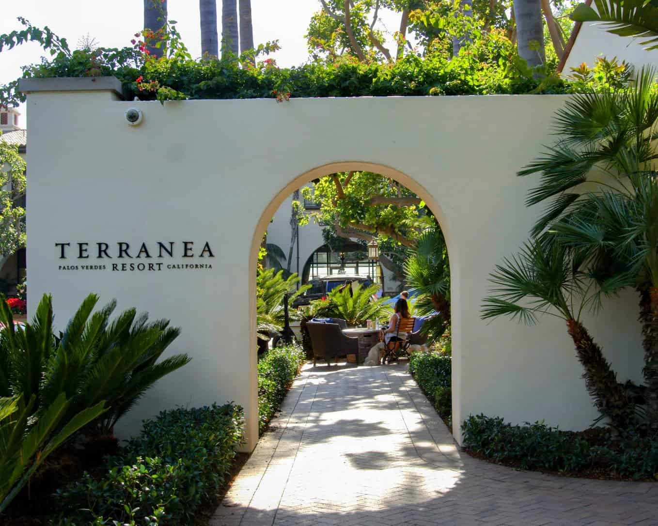 An inviting pathway at Terranea Resort in Los Angeles surrounded by lush greenery, leading through an elegant white archway to a cozy outdoor seating area. Palms and vibrant plants enhance the serene and luxurious ambiance of the resort.