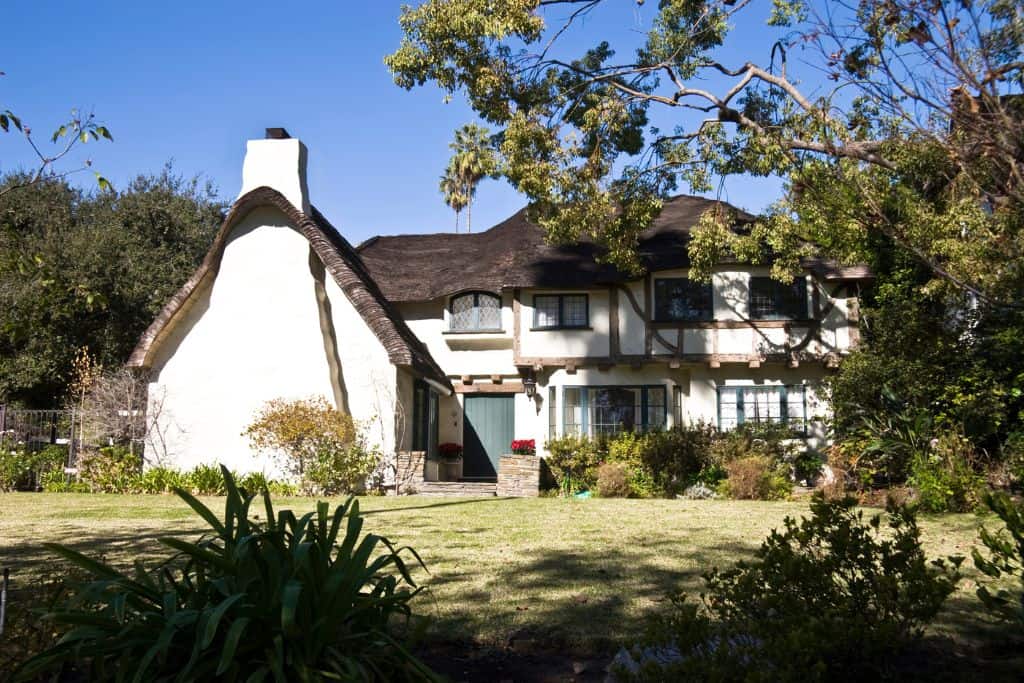 This is a photo of a charming house with a unique, wavy roof that looks a bit like a storybook cottage. It has two stories, with white walls and dark wooden trim. There's a green lawn in front with some bushes and trees around, and it's a sunny day.