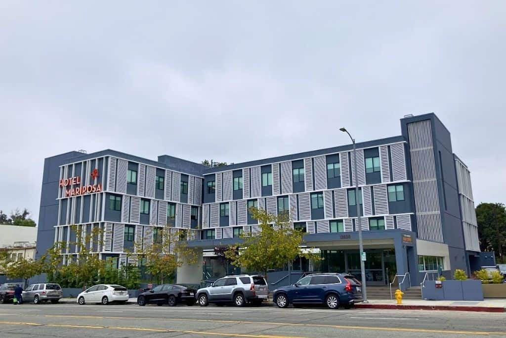 We're looking at a modern hotel building with the name Hotel Mariposa on it. The hotel has a sleek design with gray and white colors, and there are stripes on the windows. There are cars parked in front, and it looks like a cloudy day.