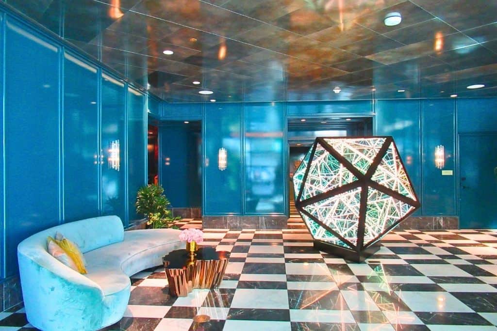 The photo depicts a luxurious interior space with teal walls and a glossy checkered floor. In the center stands a striking, illuminated geometric sculpture. A curved, plush sofa and a unique, golden table add to the opulence of the decor. The ambiance is rich and stylish, reflecting an upscale setting.
