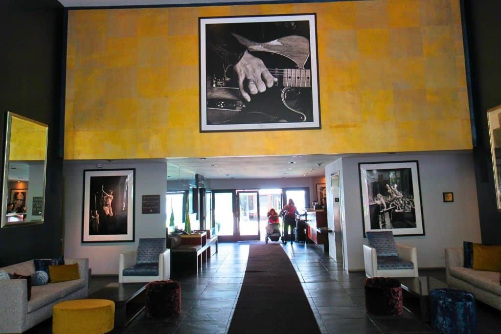 The photo features an interior lobby area with contemporary furniture and a large yellow and gold artwork on the wall. Framed black and white photographs are displayed around the room, creating an artistic atmosphere. Guests are seen in the distance, adding a sense of activity to the otherwise calm setting.