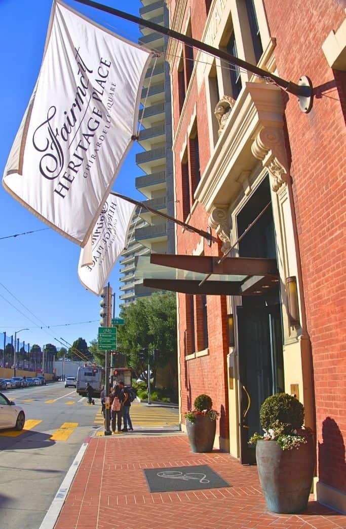 A vertical banner reading "Fairmont Heritage Place" hangs from a flagpole on a classic brick building, under a clear blue sky. Pedestrians and vehicles are visible on the street below.
