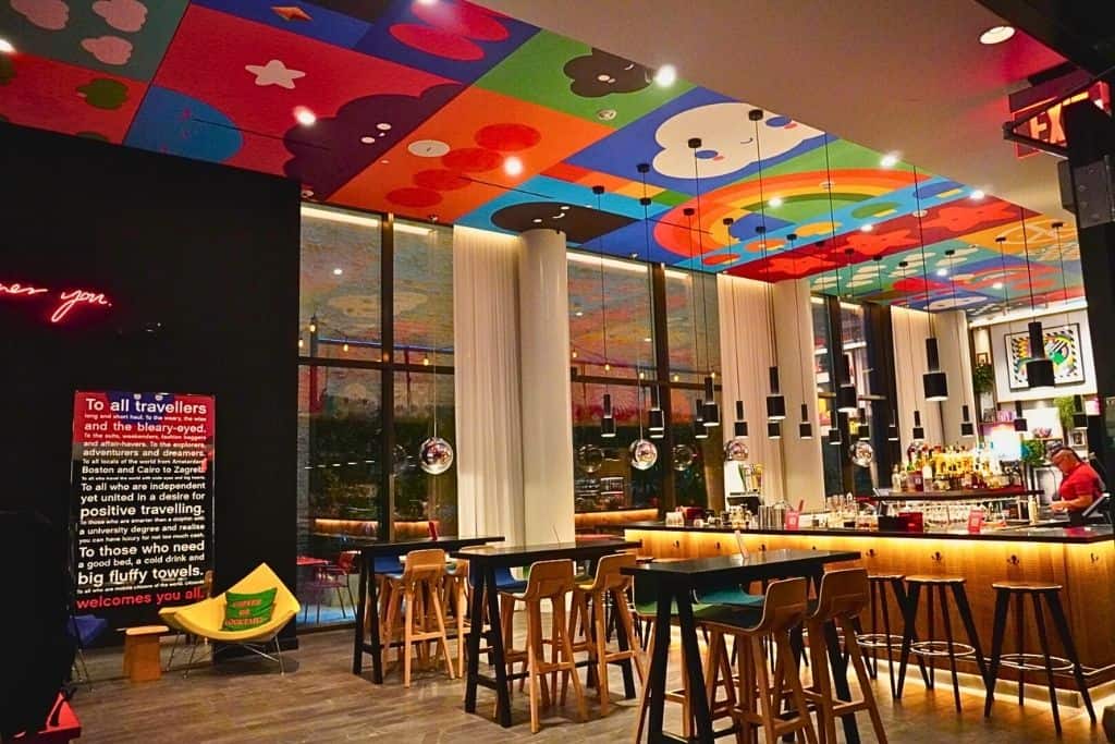 A chic bar area with a whimsically painted ceiling featuring cloud and rainbow motifs, modern furniture, and pendant lighting, inviting guests to relax and socialize.