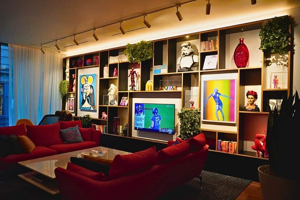 A vibrant and artsy living space with a large bookshelf filled with various colorful objects, artworks, and a life-sized Stormtrooper helmet. Red couches and a flat-screen TV create a comfortable lounge area.