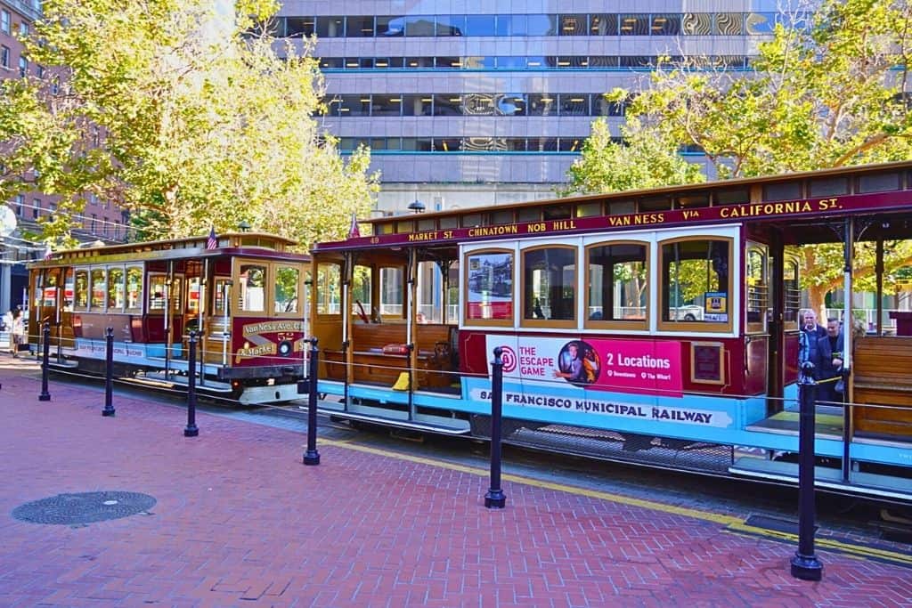 A classic San Francisco cable car stationary on the tracks, with "California St." marked as its route, set against a backdrop of city buildings and trees.